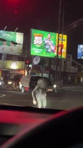 The video shows a person in a silver costume with a large, round object on their back walking across a street at night. There are several cars and motorcycles on the road, as well as illuminated billboards in the background. The person appears to be walking from the right side of the frame to the left. One billboard prominently features a man on a scooter with the text "MURAH NON STOP". Another billboard shows two people in white attire. The scene is lit by streetlights and the lights from the vehicles and billboards.