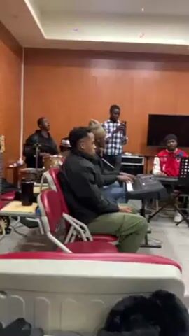 The video shows a group of young men practicing music in a room. One plays the congos, two play keyboards, and one man is recording or videoing them with a mobile phone. There is a television on the wall in the background, as well as music stands, a mixer, and a case in the foreground. The men are sitting in chairs, wearing casual clothing. The walls are wood panelled.