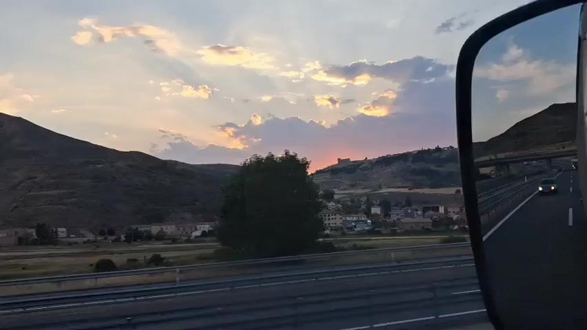 The media file captures a scenic view of a rural landscape in Medinaceli, Spain, during what appears to be sunset. The scene is viewed from a moving vehicle, indicated by the presence of a side-view mirror in the foreground, which reflects the road and a car behind.

The landscape is dominated by rolling hills, some covered in dry vegetation and others with sparse trees. A small town is nestled in the valley below, with buildings of varying sizes and colors, some appearing to be residential. On top of one of the hills, a historical structure, possibly a castle or ruins, is visible, silhouetted against the vibrant sky.

The sky is filled with dramatic clouds, illuminated by the setting sun, casting rays of light across the scene. The colors range from soft blues and purples to warm oranges and yellows, creating a serene and picturesque atmosphere. The time of day is clearly sunset, as suggested by the lighting and the colors of the sky.

The main activity implied is travel, given the perspective from a vehicle and the presence of a highway with lane markings. There are no people clearly visible in the scene, but the car in the mirror suggests other vehicles are present. The overall mood evoked is one of peaceful travel and appreciation of natural beauty. No visible text is present in the media file.