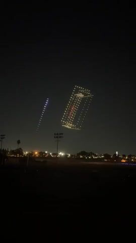 The video displays a nighttime aerial drone show above a dark, open field with a line of trees and distant city lights in the background. The main focus is a large formation of drones creating a glowing, three-dimensional shape resembling an open rectangular box with diagonal bracing. The drones are illuminated with bright yellow and white lights, outlining the structure. Within the box, some drones are lit with red lights. To the left of this larger formation, a smaller, vertical string of purple and white lights, also formed by drones, ascends into the sky. A tall, dark silhouette of a utility pole with lights is visible in the foreground, slightly to the left of the center. The sky is dark and clear, suggesting a cloudless night. The drones are clearly organized and moving in a coordinated pattern, indicative of a programmed light show.