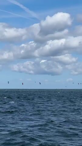The video shows a wide expanse of choppy, dark blue water under a bright blue sky with fluffy white clouds. The horizon is visible, and in the distance, there are numerous kitesurfers, identifiable by their colorful kites. The kites are spread out across the water, some closer to the viewer and some further away, indicating an active kitesurfing area. The lighting suggests it is daytime.