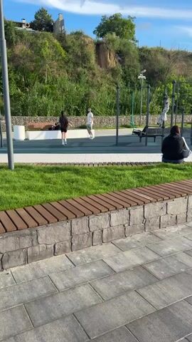 The video captures an outdoor park or public exercise area on a clear, bright day. In the foreground, grey rectangular paving stones lead up to a low wall constructed from rough-hewn grey stones, topped with wooden slats, bordering a strip of vibrant green grass.

Beyond the grass, a blue-surfaced exercise zone is visible, equipped with various metal workout structures like pull-up bars. On this surface, two individuals are engaged in physical activity. One person, dressed in dark attire, is moving around, possibly walking or jogging. Another, wearing a white t-shirt and dark shorts, is performing squats or lunges. Further to the right on the exercise surface, a third person is partially seen near some equipment.

In the mid-ground, to the right, a person in a dark hoodie is seated on a stone bench, facing away from the camera. Tall metal poles, likely for lighting or supporting structures, are interspersed throughout the scene.

The background features a prominent hillside covered in dense green foliage and trees, with some buildings visible atop the hill under a bright sky with scattered clouds. The overall scene suggests a pleasant environment for recreation and exercise. No discernible text is visible within the frame.