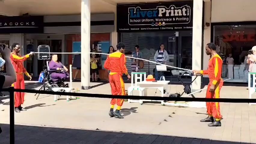 The video depicts a street performance in front of shops. Three individuals dressed in matching red and yellow outfits are the main performers. Two of them are holding a long, white rope taut between them, and the third performer is actively jumping over the rope as it is being swung. The jumping performer is in mid-air, with their legs bent, suggesting a dynamic movement. To the left, another individual is holding a phone, seemingly recording the performance. In the background, a person in a wheelchair is seated, observing the show. Behind the performers, there is a table with some items on it, and a keyboard or synthesizer. The environment appears to be a pedestrianized area, possibly a shopping precinct, with store fronts visible, including one labeled "Liver Print". The lighting suggests it is daytime, with bright sunlight casting shadows. Several other bystanders are visible in the background, some watching the performance.