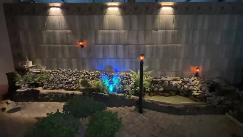The video shows an outdoor backyard area at night. The main focus is a decorative wall with large grey rectangular tiles, illuminated by several uplights at the top. Below the wall is a rock garden with a small pond and lush greenery. Colorful lights, cycling through blue, red, and green, illuminate a waterfall feature within the rock garden. Two small orange outdoor sconces are mounted on the wall. A single black lamppost with an orange light fixture stands in the foreground, in front of the rock garden. The ground is paved with brick-like stones. In the lower left corner, a person's feet and legs can be seen, along with a small table and chair. In the lower right corner, some green bushes are visible. The lighting creates a dramatic and serene atmosphere.