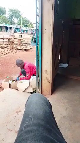 The video shows a person sitting and facing forward, with their gray jeans visible in the foreground. In the midground, a child wearing a red shirt is kneeling beside a partially open cardboard box, looking into it. A man wearing a plaid shirt and carrying multiple bags walks in the background, from left to right, behind a blue gate. People and motorcycles are visible in the distance. The scene appears to be outside a wooden structure, possibly a shop or shed, with a reddish-brown dirt ground.