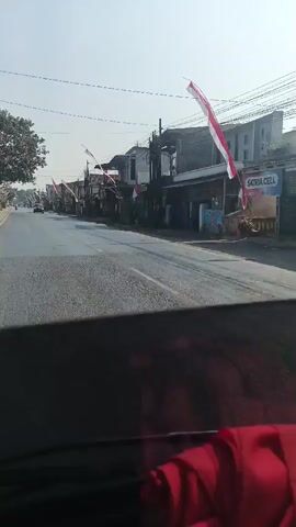 The video shows a view from inside a vehicle looking out at a street lined with buildings and trees. Indonesian flags are visible, indicating a possible national celebration or event. Several cars and motorcycles are traveling on the road. The sky is clear and the weather appears to be sunny. The overall scene depicts a typical street view in what appears to be a residential or commercial area.