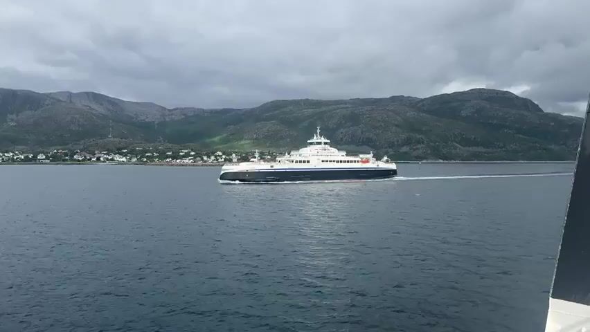 A wide shot shows a large white and dark blue ferry traveling across a calm, dark blue body of water. The ferry is positioned in the center of the frame, moving from left to right, and leaving a wake behind it. In the background, a line of green, rocky mountains dominates the horizon. A small town with houses and buildings is visible along the shoreline at the base of the mountains. The sky is overcast with heavy, gray clouds. The lower right corner of the frame shows the edge of another vessel, likely the one from which the video is being recorded. The overall scene is somber due to the cloudy weather.