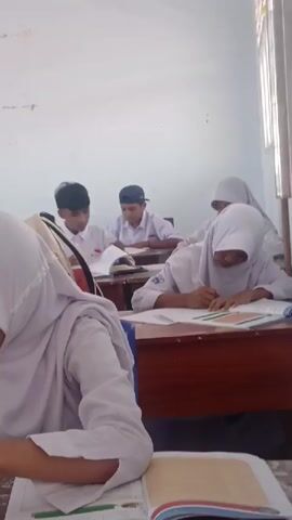 The video shows a classroom scene with several students sitting at desks and engaged in writing. The students are wearing white uniforms, some with headscarves. The desks are wooden, and books and papers are spread out on them. The lighting suggests it is daytime, with natural light coming from a window. The overall atmosphere appears to be one of focused study.