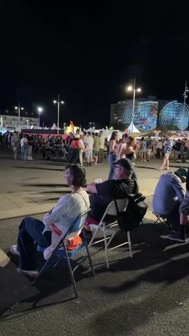 The video shows an outdoor nighttime scene, likely a festival or event. In the foreground, two people are seated in chairs, facing away from the camera. The woman on the left wears a light-colored top and dark pants. The man on the right wears a black t-shirt and a baseball cap, with a backpack next to him. The background is a crowded open area with several tents, stage structures, and a Ferris wheel or similar illuminated attraction. People are milling about, some standing and some walking. Lights from the event illuminate the scene, and the sky is dark. There is a general atmosphere of a public gathering at night.