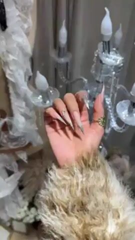 A close-up shot shows a person's hand with extremely long, stiletto-shaped fingernails. The nails are decorated with a gradient design, transitioning from a light pink or nude color at the base to a dark grey or black at the tips. One nail features small, silver embellishments. The person is wearing several gold rings, including one with an intricate, flower-like design. The hand is positioned against a backdrop that includes a decorative candelabra with faux candle bulbs and a sheer, white, frilly material. The person's arm is clad in a fuzzy, light-colored jacket. The lighting is soft, creating a slightly hazy and somewhat ethereal atmosphere. The camera remains mostly still, with minor adjustments to keep the hand in focus.