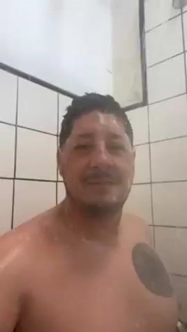 A shirtless man with a tattoo on his left chest is taking a shower. He has dark hair and a mustache, and is facing the camera. The shower stall is tiled with white squares, and water is running from the shower head. The man raises his right hand to his head.
