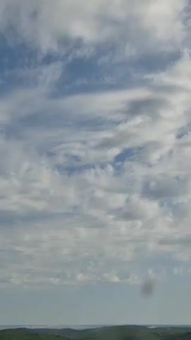 The video shows a time-lapse of clouds moving across a blue sky with scattered white clouds. The landscape below is green and appears to be rolling hills with trees. There is a dark, blurry spot in the middle of the frame which may be a lens artifact or something in the environment. The time stamp indicates the video lasts approximately 5 seconds.