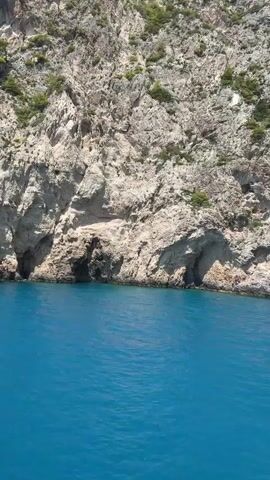 A boat is moving past a steep, rocky cliff face that rises from the turquoise sea. The cliff is textured with various shades of gray and white rock, with patches of green vegetation growing in crevices. There is a dark opening in the rock face near the waterline, suggesting a cave or an inlet. The water is clear and calm, with gentle ripples on the surface. The sky is visible at the top of the frame, appearing bright and sunny. The video seems to be shot from the perspective of someone on the boat.
