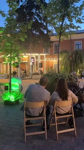 A group of people are seated on chairs outside a brick building at dusk. Two people are in the foreground, facing away from the camera, looking towards a group of people gathered around a stage or performance area. The building has a covered outdoor seating area with lights and a sign that is partially visible. There are trees and plants in the scene, some of which are illuminated with green lights. A stroller is visible to the right of the two people in the foreground. The overall atmosphere appears to be relaxed and social, with an outdoor performance or event taking place.