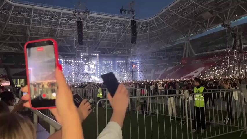 The video captures a concert or large event taking place in a stadium at dusk. A large crowd of people is gathered, many holding up their phones with flashlights illuminated, creating a sea of lights. A stage with multiple light structures and screens is visible in the distance, where a performer is likely present, though not clearly discernible. Two individuals in the foreground are holding up smartphones to record the event. One phone, in a red case, is held vertically, while the other, a dark-colored phone, is held horizontally. Security personnel in bright yellow vests are positioned among the crowd behind a metal fence. The stadium itself has a distinctive roof structure and seating areas filled with spectators. The overall atmosphere appears to be one of excitement and engagement from the audience.
