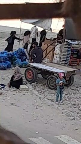 A group of people are gathered outdoors in a dusty, sandy environment, likely a marketplace. In the foreground, a person wearing a headscarf is seated on the ground next to some items. To their right, a child in a pink hat stands. Behind them, a man is seated on a cart with two wheels, facing away from the camera. A few other individuals are visible in the background, some standing and one seated near a stall covered by a white awning. Blue bags are piled up near the cart. The ground is covered in dirt and small stones.