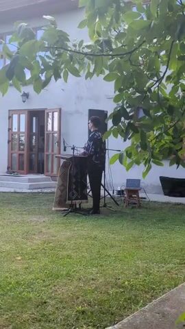 A person wearing a black and white patterned shirt and black pants stands behind a wooden stand adorned with a patterned rug. They appear to be playing an instrument. To their right is a speaker and a small laptop on a stool. A woman in a red dress walks from the open doors of a building into a grassy yard. The scene is outdoors with trees and leaves in the foreground.
