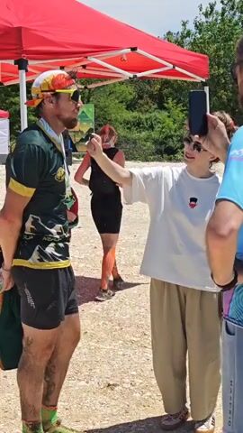 A man wearing a green and yellow sports jersey, black shorts, and a colorful cap stands facing right, his body covered in mud stains. He holds a plastic water bottle. A woman in a white t-shirt and light brown pants stands facing him, holding a dark smartphone to film him. Behind them, another woman in a black top and shorts drinks from a bottle. Two red tents are visible in the background, with greenery beyond. The ground is light brown and dusty.