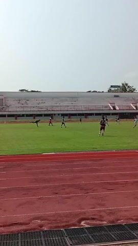 The video shows a soccer game being played on a field with a red running track around it. Several players wearing different colored jerseys are scattered across the green field, actively participating in the game. In the background, there is a stadium with white and red seating. A tall light pole is visible in the distance. The sky is bright and clear. The camera is positioned on the edge of the running track, capturing the action on the field.