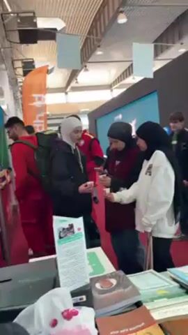 The video shows an indoor event, likely a fair or exhibition, with various people walking around. Three women wearing hijabs are standing at a table covered with books and informational materials, looking at a smartphone. Other people are walking in the background, including men in casual attire and someone in a red jacket. A booth with a large screen and a black backdrop with light fixtures is visible behind them. The floor is covered with red carpeting. The building has a high ceiling with exposed beams and numerous lights.