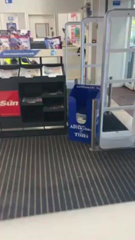 A person in a mobility scooter is seen passing through a security gate at the entrance of a store. The individual is wearing a bright yellow safety jacket and dark pants. The security gate is white and has several horizontal bars. To the left of the gate, there is a newspaper stand featuring publications like "The Sun," along with various magazines. A blue dispenser for "New Milton Advertiser & Times" newspapers sits next to the gate. The floor is covered with a striped gray and white mat. The gate alarm sounds as the person passes through. The background includes a blurred view of the store's interior, with shelves and doors visible.
