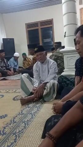This is a video captured indoors, likely in a community hall or large room. Several men are seated on the floor, which is covered with mats and rugs. Many of the men are wearing traditional Islamic attire, including peci caps and sarongs. In the foreground, an older man with a white shirt and striped sarong is seated, appearing to be the focal point of the gathering. Behind him, other men are seated in rows. In the background, there are chairs stacked up, a large speaker, and a window. A white pillar is also visible on the right side of the frame. The lighting appears to be artificial, suggesting the event is taking place in the evening or a dimly lit interior. The overall atmosphere seems to be one of a religious gathering or a community meeting.