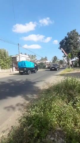 A black SUV with a blue tarp covers on its roof drives down a road. The SUV is followed by another vehicle which is partially obscured by the SUV. A person rides a motorcycle down the road towards the SUV. Green vegetation is on the right side of the road. A building with a red door is on the left side of the road. The sky is blue with some white clouds.