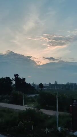 This is a vertical, medium shot video of a cloudy sky at dusk. The sun is peeking through the clouds on the left side of the frame, casting a soft glow. Below the sky is a landscape with trees, buildings, and a parking lot full of cars. There are also some power lines and streetlights visible. The overall mood of the video is peaceful and serene.