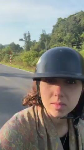 A woman wearing a black helmet rides a motorcycle down an empty road with lush green vegetation on either side. The sky is bright and clear. A vehicle drives down the road in the opposite direction. The woman seems to be filming herself as she rides.