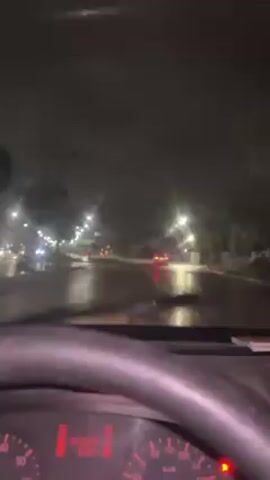 The video shows a dashboard view from inside a car driving on a rainy night. Streetlights are visible on either side of the road, illuminating the wet pavement. Cars with red taillights are visible ahead. The car's dashboard shows the speedometer and other instrument panel indicators. The windshield wipers are in use. The camera appears to be stabilized, as the view is relatively steady despite the moving car.