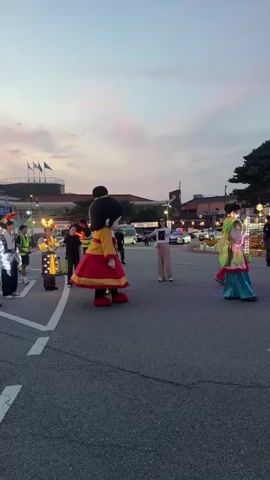 This is a vertical, low-angle, outdoor shot of what appears to be a parade or street performance at dusk. The sky is a dusky pink and orange, indicating the sun has recently set or is about to. In the foreground, the asphalt road has white lane markings. To the left, there is a group of people dressed in elaborately decorated, illuminated costumes, some holding what look like large lanterns. They are standing in a line and appear to be part of a performance. Further back and slightly to the right are two large mascot-like characters. One, on the left, is a cartoonish figure with a turquoise cap, green shirt, and yellow pants. The other, on the right, is a character in a red and yellow outfit with a large head and black hair. A woman in white is standing behind the red and yellow mascot. In the background, there is a building with flags flying, streetlights, cars, and trees. The overall impression is a festive event with performers, costumes, and public gathering.