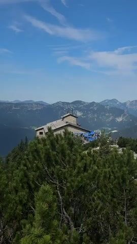 The video pans across a mountainous landscape. In the foreground, lush green pine trees are visible. In the mid-ground, a stone building with a brown roof sits on a hilltop. Blue umbrellas are spread out on a patio area next to the building. Beyond the building, rolling green hills transition into darker, more rugged mountains in the distance. The sky is a clear blue with a few wispy white clouds. The scene appears to be a scenic overlook or a mountain lodge in a natural environment.