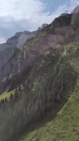 This video shows a view from a cable car ascending a steep, tree-covered mountain. The cable car's wires and tower are visible at the top of the frame. The mountainside is rugged with rocky outcrops and patches of green grass and dense evergreen trees. The sky is cloudy.