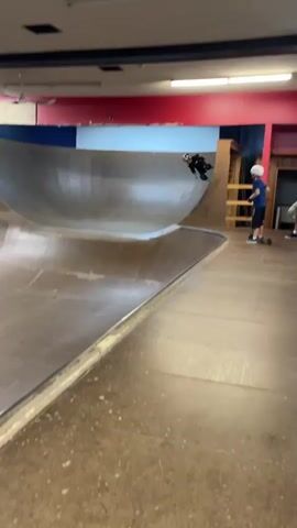 A skateboarder is performing a trick in a skatepark. The skateboarder is wearing protective gear, including a helmet and knee pads. The skatepark features a large, curved ramp. Another person on a scooter is standing in the background. The environment appears to be indoors, with wooden walls and red and blue accents on the ceiling.