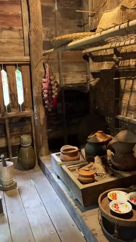 The video pans across the interior of a rustic kitchen. The walls and floor are made of wood, giving the space a traditional feel. In the foreground, a wooden mortar and pestle sits on the floor next to a large ceramic jar. A bundle of garlic and red chilies hangs from a hook on the wall. In the center of the frame is a traditional cooking hearth with clay pots and a large black cauldron placed over burning embers. Shelves above the hearth hold more kitchenware, including woven baskets and additional pots. A woven tray with colorful plates is visible on the right side of the frame. The lighting is dim, suggesting an indoor setting, possibly a museum exhibit or a preserved historical dwelling. The overall impression is one of a simple, functional kitchen from a past era.