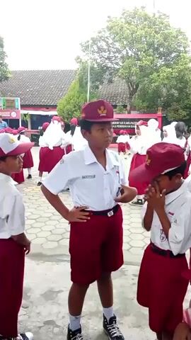 A group of young students, all wearing matching school uniforms consisting of white shirts, maroon shorts or skirts, and maroon caps, are gathered outdoors on a paved surface. The boy in the center, facing the camera, is wearing a white short-sleeved shirt, maroon shorts, black and white sneakers, and a maroon cap. He has his left hand on his hip. To his right, another boy in a similar uniform is facing him, with his hands clasped in front of him. Other students, mostly facing away from the camera, are visible in the background. They are also dressed in the same school uniforms. Behind the students, there are school buildings with red roofs and walls, and a tall antenna structure can be seen in the distance. Trees with green foliage are present in the background. The overall scene appears to be a schoolyard, possibly during a break or an assembly.