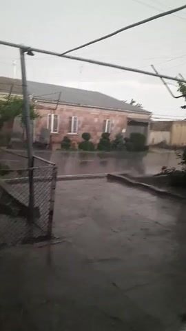 A cloudy, rainy day is depicted from a fenced-in yard looking towards a residential street. Raindrops can be seen hitting the wet pavement and creating ripples. A single-story house with a red-tiled roof and light pink stone walls is visible across the street, along with a manicured garden in front of it. The sky is overcast and gray, and power lines stretch across the frame. There are no people visible in the video. The scene conveys a sense of quiet solitude during a downpour.