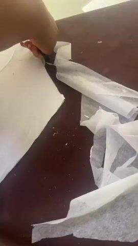 A close-up view shows a person's dark-skinned hands actively working with white fabric on a brown table. The hands are seen manipulating the lightweight white material, smoothing it, and cutting it with a pair of black-handled scissors that rest on the table. Several scraps of the white fabric are also visible around the workspace. In the background, portions of a metal-topped work surface or sewing machine, a yellow measuring tape, and some red objects are faintly discernible. The activity appears to be fabric preparation, possibly cutting patterns or interfacing for a sewing project, within an indoor workshop or crafting setting. No text is visible.