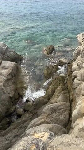 The video shows a close-up, overhead view of clear, turquoise water gently lapping against a rocky shoreline. The rocks are various shades of gray and green, with some appearing mossy. Small waves create white foam as they break against the rocks. The water is shallow enough to see the rocky bottom and some small stones. The sky appears overcast, contributing to the muted tones of the scene. There are no people or man-made objects visible. The camera angle is stable, suggesting it might be mounted on a tripod or held by someone standing still.