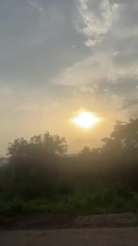 The video captures a serene sunset over a rural landscape in Umuoye, Nigeria. The bright, low sun is visible through scattered clouds, casting a warm glow over the scene. Lush green vegetation, including trees and dense undergrowth, dominates the foreground and midground, with hills or mountains discernible in the distant haze. The footage is taken from a moving vehicle, and at one point, the rear and side of a silver sedan become visible, with the sun's rays reflecting off its surface. The car's license plate, which reads "17M ABIA" and "GWA-315CY," is clearly visible.