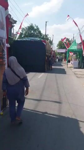 A low-angle, medium shot shows a street scene with several people and a large, dark-covered structure on the right side of the road. The structure appears to be a temporary stall or display, decorated with what looks like straw or thatch on top, and adorned with Indonesian flags, which are red and white. Several people are visible near the structure, some appearing to be vendors or organizers, and others are walking past or observing. A person in the foreground on the left, wearing a light purple headscarf and a light blue top, is walking away from the camera. The street is paved with asphalt and has white lane markings. In the background, there is a black SUV, more flags, and green tents. The sky is blue with some clouds, and trees and buildings are visible along the sides of the road. The overall impression is of a festive or event-related atmosphere, possibly a market or a parade setup.