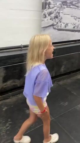 A young girl with blonde hair and fair skin, wearing a light purple crop top over a white and yellow patterned shirt and white shoes, walks and dances in a museum or exhibition space. The girl is filmed from the side and then from the front. She laughs and appears to be enjoying herself. Behind her is a black wall with a large black and white mural or display of historical images depicting a cityscape with boats and people. The floor is dark and appears to be tiled. The video ends with a wider shot of the girl moving down a long corridor with similar displays on the walls.