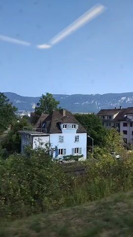 The video captures a view from a moving vehicle, likely a train, looking out at a residential area with houses and lush greenery. The sky is a clear blue with faint white streaks, possibly from clouds or light reflections. In the foreground, there is blurred foliage, indicating the camera's movement. Several houses are visible, ranging from a light blue two-story home to larger apartment buildings with balconies and reddish roofs. A white car is parked on a driveway. In the background, distant mountains with white peaks are visible under the blue sky. The overall impression is a serene, suburban landscape on a bright day.