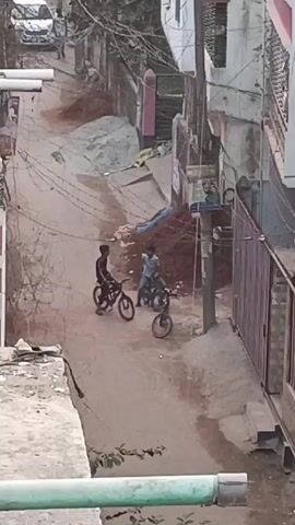 The video shows an elevated view of a narrow street in what appears to be a residential area. The street is unpaved and appears to be made of dirt. There are three boys with bicycles on the street; two are on their bikes, and one is walking beside them. The boy on the left is wearing a dark shirt, while the boy on the right is wearing a light-colored shirt. One additional individual, wearing dark clothing and carrying an object, is walking away from the group in the distance. Buildings line both sides of the street, with fences and walls visible. There are electrical wires overhead, and piles of dirt or building materials are scattered along the street. A parked car is visible near the top of the scene. The footage is slightly shaky, suggesting it was hand-held.