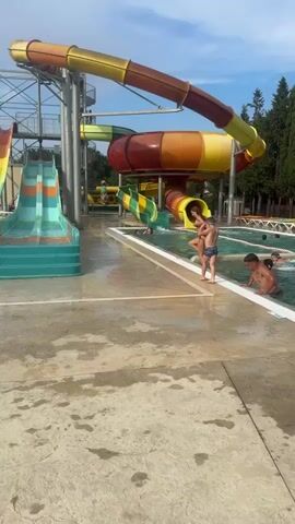 The video shows a water park on a sunny day. There are several water slides visible, including a large, winding yellow and red slide and two smaller, straighter slides with green and orange colors. In the foreground, a person is walking on the wet concrete surrounding the pool area. In the middle ground, a man is standing in shallow water and a child is being held by another person. Further back, there are lounge chairs and a covered area, as well as other people enjoying the water. The overall atmosphere appears to be lively and recreational.