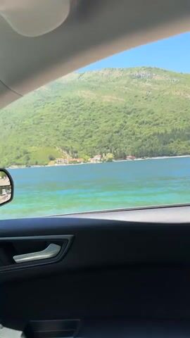 The video captures a view from inside a car, looking out the passenger window. The window frames a scenic landscape dominated by a large, green, tree-covered mountain that slopes down towards a bright blue body of water. Along the shoreline at the base of the mountain, a scattering of buildings, appearing to be houses or small structures, are visible. The water itself is clear and turquoise, with some ripples on its surface indicating a gentle breeze or movement. The car's interior, specifically the dark-colored door panel with a silver door handle, is in the foreground, partially obscuring the view. The sky is a clear, vibrant blue. The camera appears to be moving slowly, as the scenery outside shifts subtly.