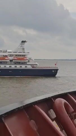 The video shows a ship at sea passing by another ship. The passing ship is named Seadream Yacht Club. The sky is overcast and gray. The water is murky brown. The ship filming the video has a red railing.