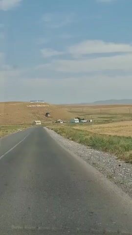 A first-person perspective video captures a paved road stretching through a dry, grassy landscape under a partly cloudy blue sky. On the right side of the road, there's a strip of gravel and dry vegetation. In the distance, a gentle hill is visible, topped with large white letters that spell out a word or phrase. Several small, simple houses are scattered across the landscape, some near the road and others closer to the hill. The terrain appears arid and somewhat barren, with fields of dry, yellowish crops extending towards the horizon. The overall impression is of a rural, possibly remote, location on a sunny day.