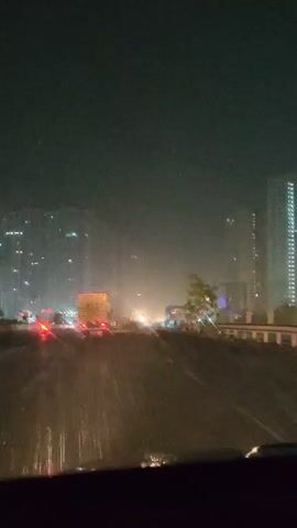 A night scene with a view through a wet windshield on a highway. In the distance, there are tall buildings illuminated with lights. The highway is filled with vehicles, their headlights and taillights creating streaks of light. Some vehicles appear to be stopped, suggesting traffic congestion. The overall atmosphere is dark and somewhat hazy, possibly due to rain or mist.
