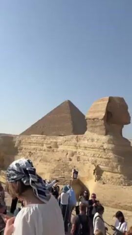 The video shows the Great Sphinx of Giza and a pyramid in the background, under a clear blue sky. There are many tourists walking around and taking pictures. The camera pans to the right, revealing more of the desert landscape and stone structures. Some individuals are wearing casual clothing and hats.